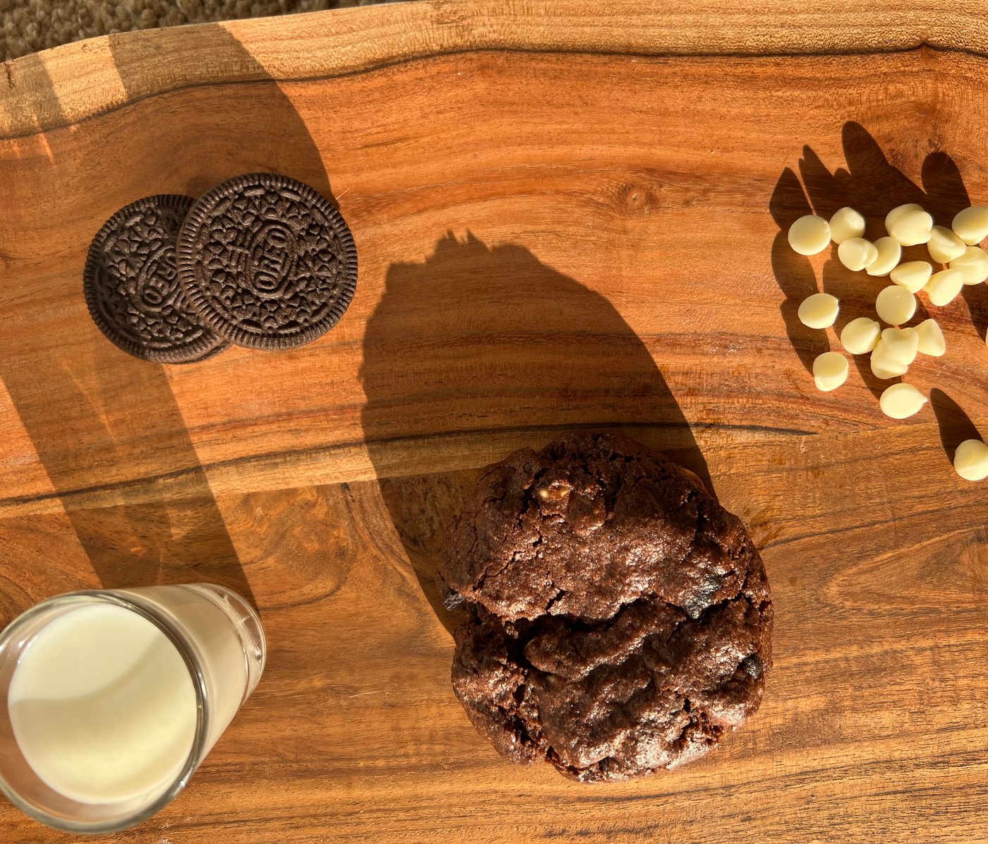 A double chocolate cookie on a wooden board with a glass of milk, Oreos and white chocolate chips